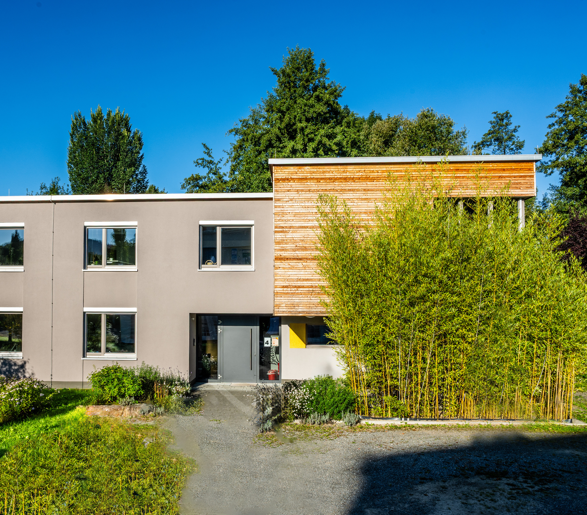 Die zurückgesetzte Eingangstür ist von Fenstern umgeben. Dieses Muster wird durch den Holzkubus unterbrochen, der rechts anschließt.
