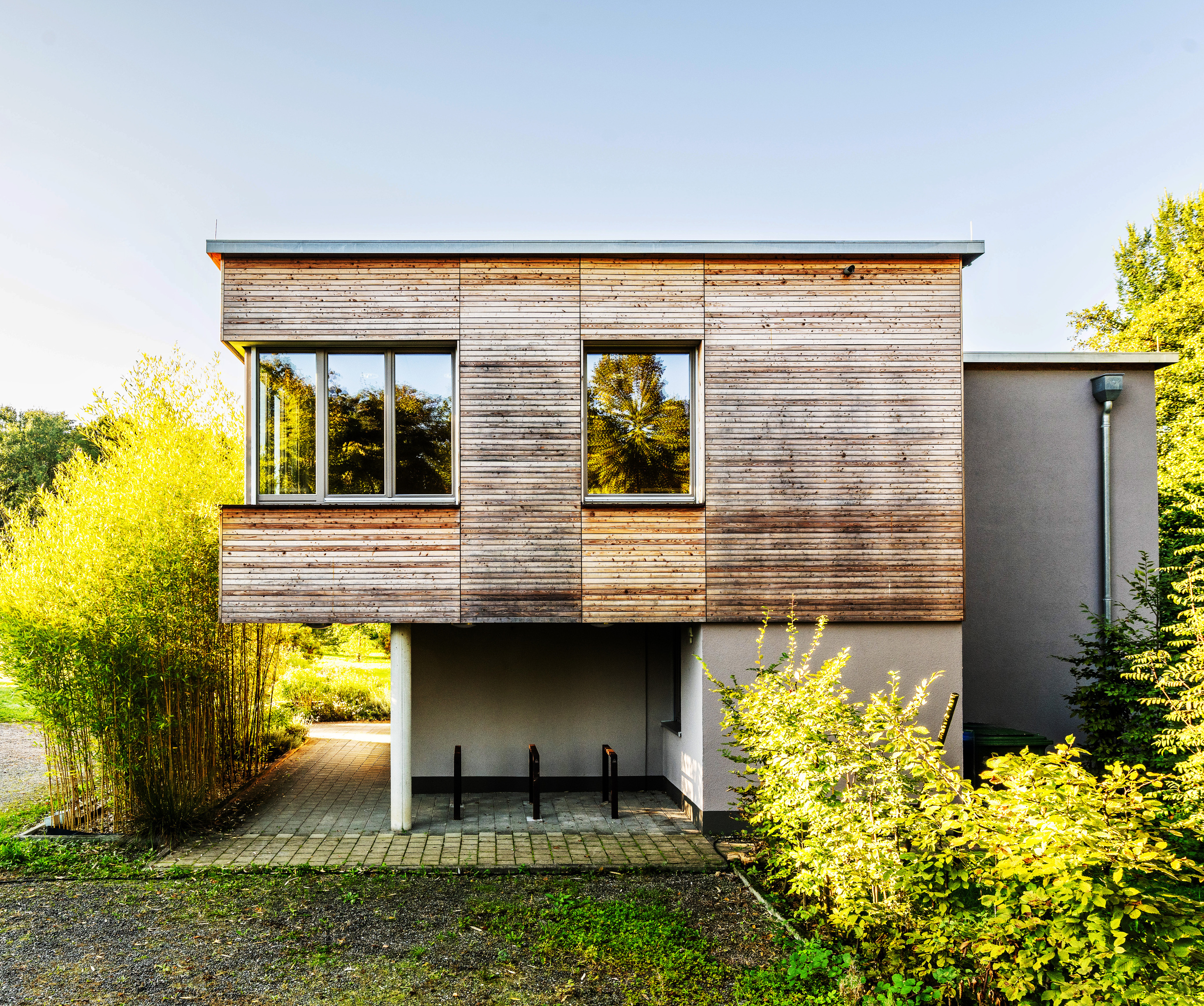 Aussenansicht des Holzkubus im ersten Stock. Er ist mit horizontalen Holzlatten verkleidet und bildet einen regengeschützten Unterstand für Fahrräder.