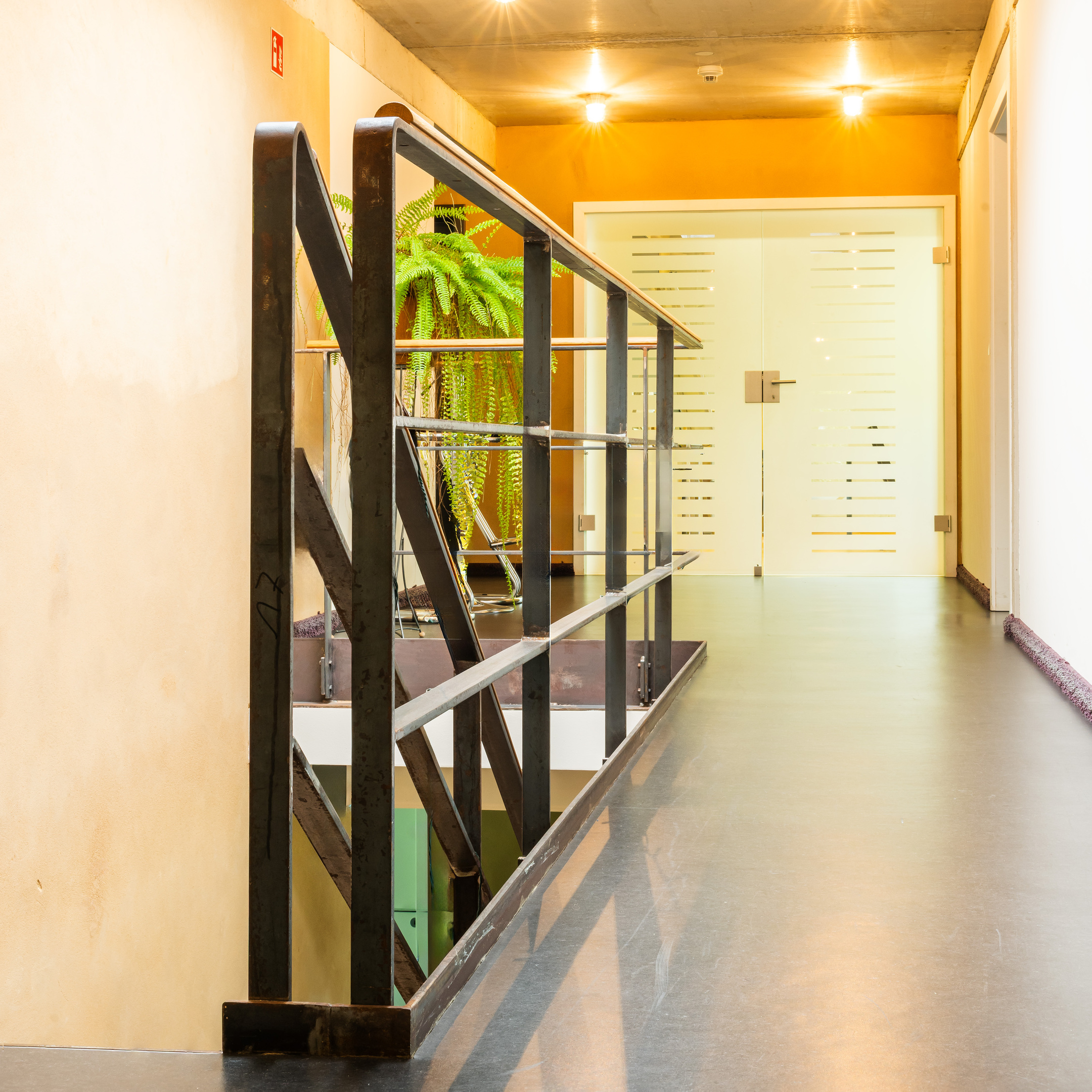 Flur im Obergeschoss. Das Treppengeländer ist aus gebogenen Stahlbändern mit einer Holzauflage. Im Hintergrund eine Doppelflügeltür aus Milchglas.