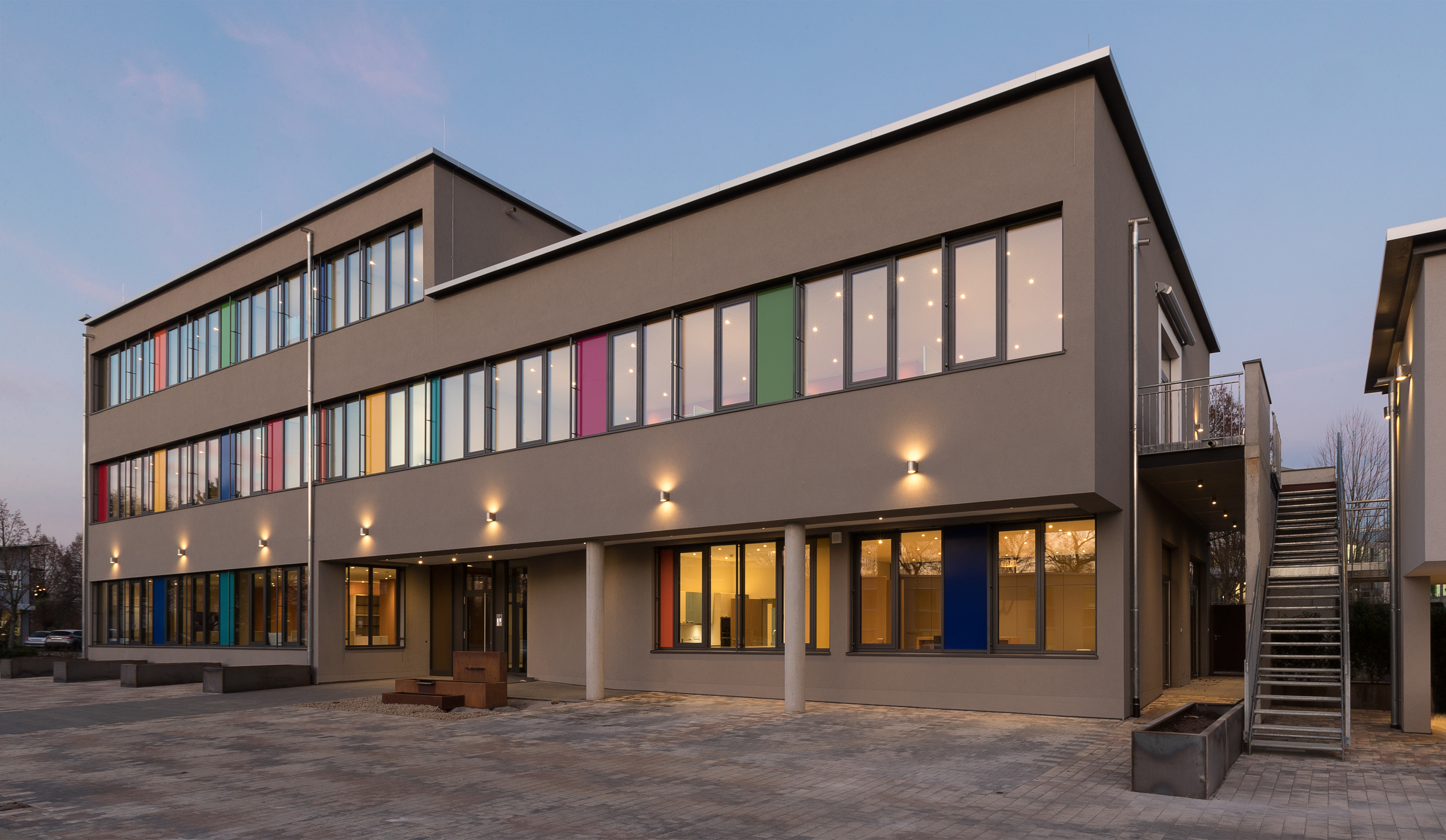 Dreistöckiges Gebäude. Das oberste Stockwerk geht über die halbe Breite. Zurückgesetzter Eingangsbereich. Durchgehende Fensterbänder, die mit farbigen Platten durchbrochen sind.