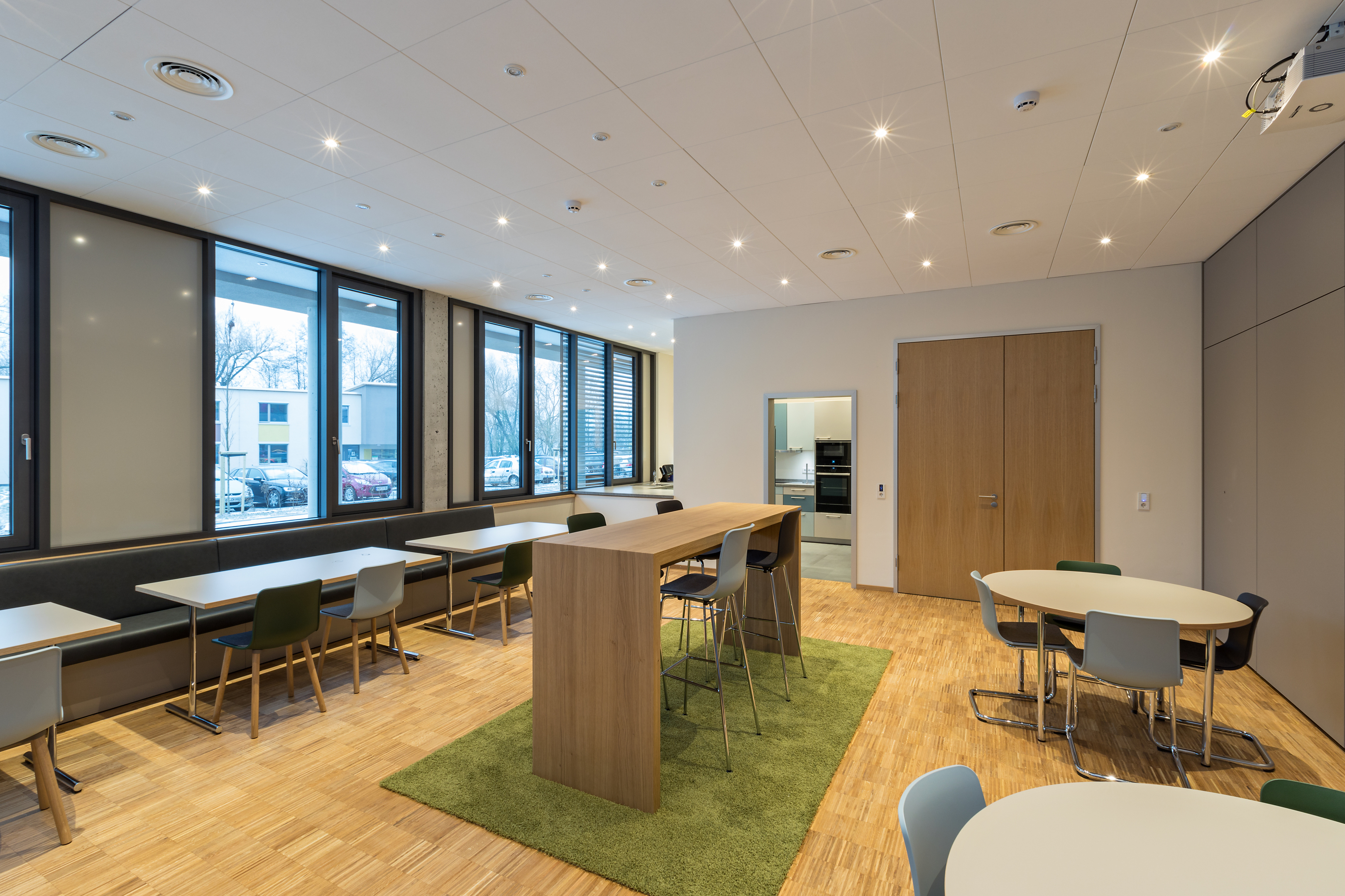 Raum mit einem grünen, dicken Teppich in der Mitte. Sechs runde und rechteckige Tische sind im Raum verteilt. Entlang der Fensterfront eine Sitzbank.