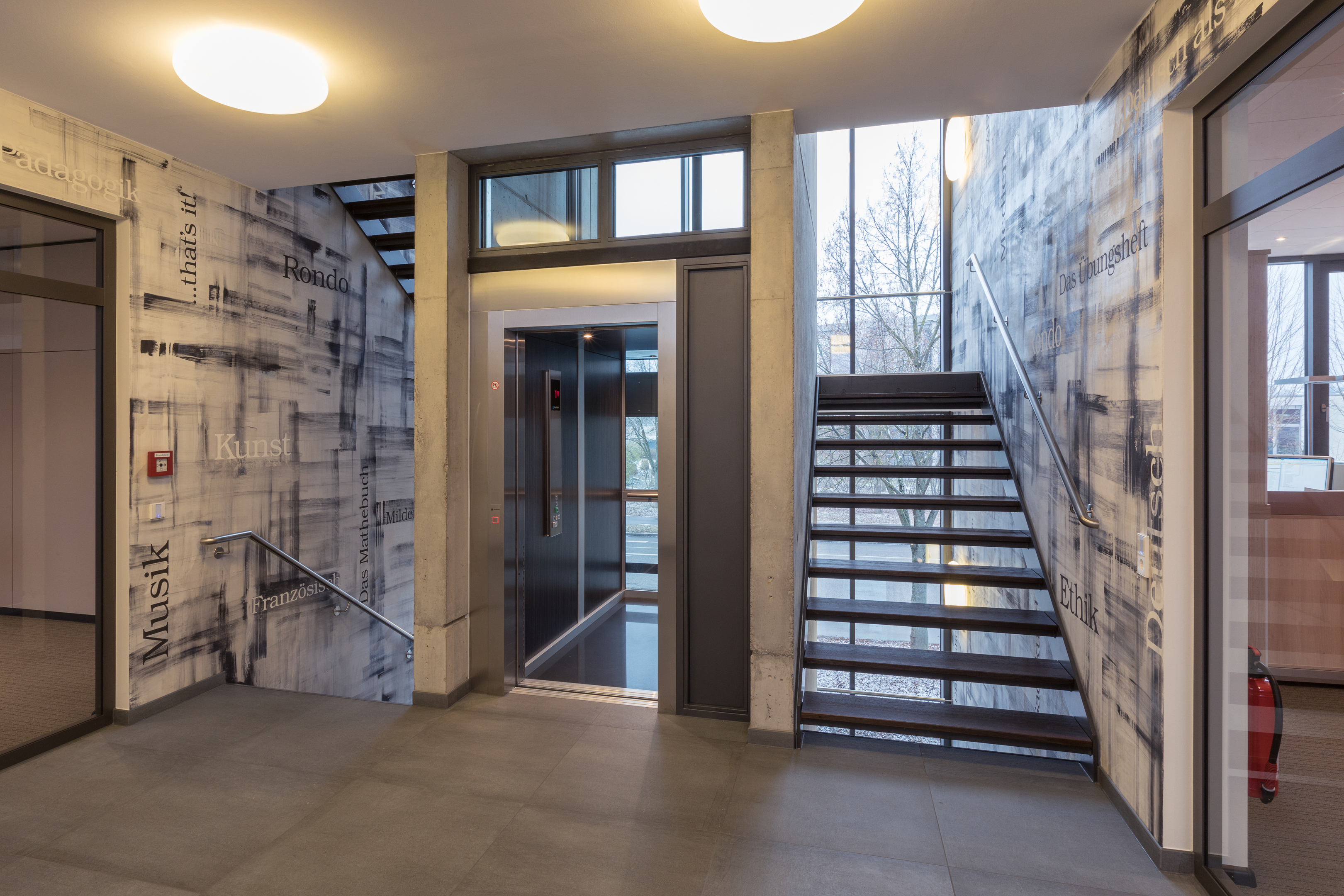 Treppenhaus mit Aufzug und Fensterfront. Die Wände sind in Graustufen, die an Druckerschwärze erinnern und orthogonal angeordneten Worten bemalt.