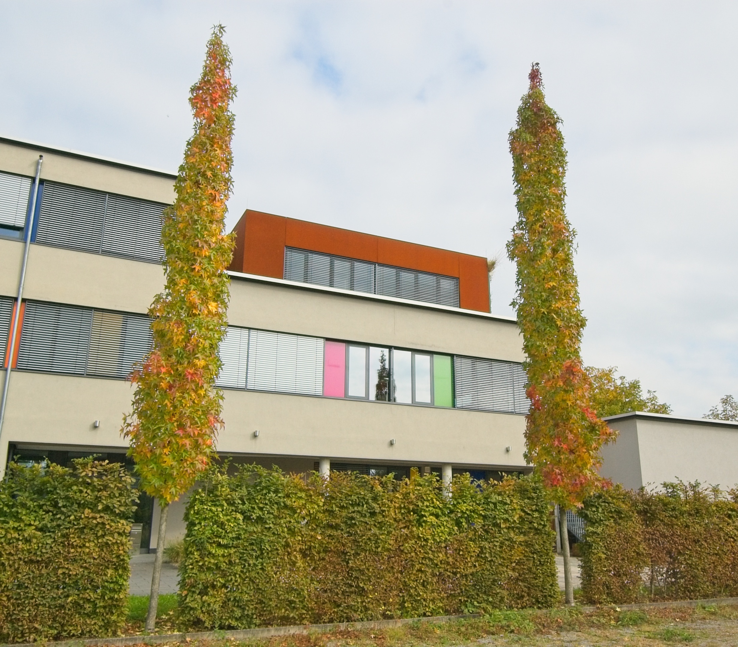 Dreistöckiges Bürogebäude. Im dritten Stock ist auf der Dachterrasse der Kubus aus Cortenstahl mit seinem breiten Fensterband zu sehen.