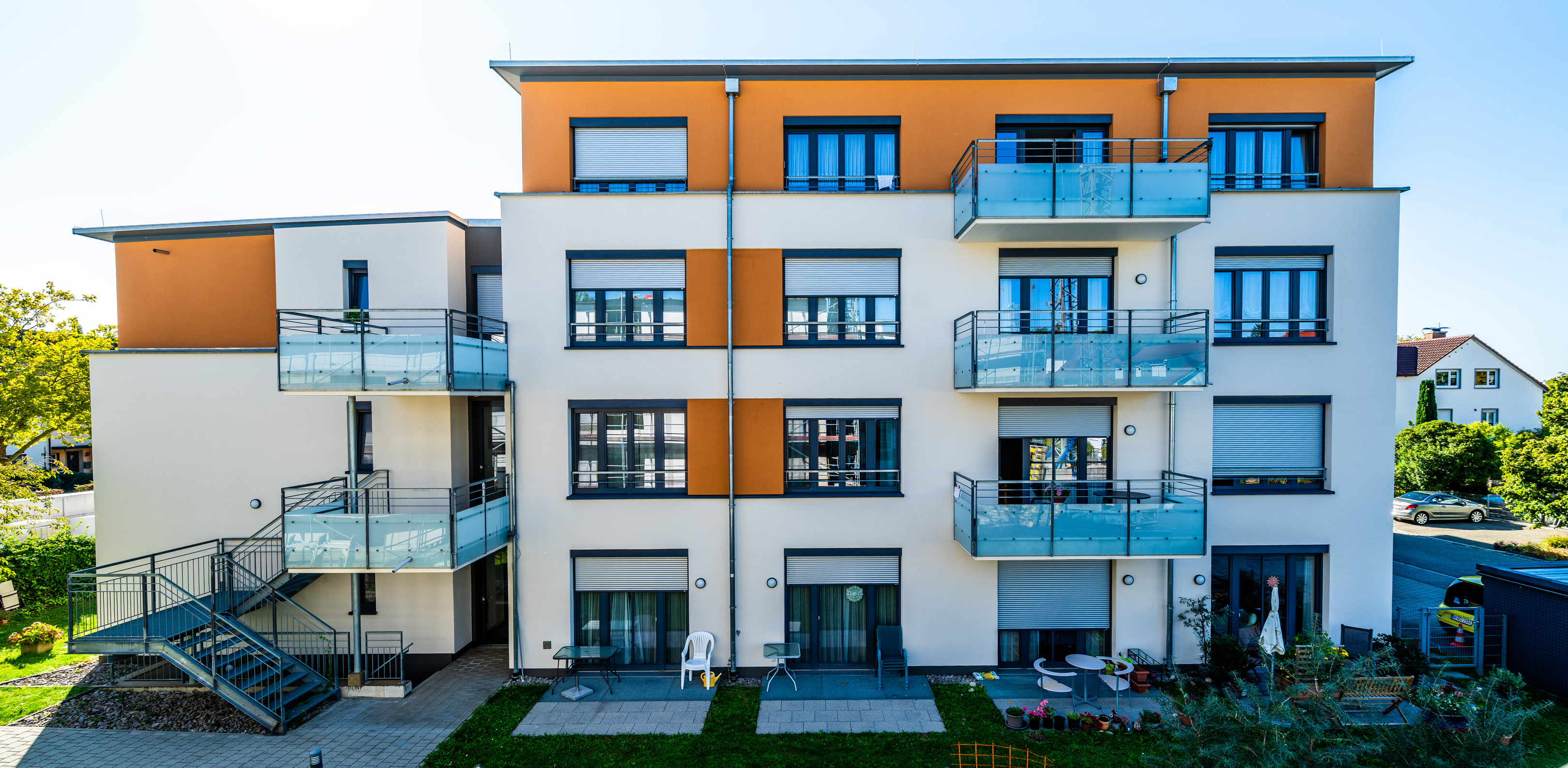 Vierstöckiger Wohnteil. Im Ergeschoss Gartensitzplätze, oben großzügige Balkone. Oberstes Stockwerk in Orange akzentuiert und leicht zurückgesetzt.