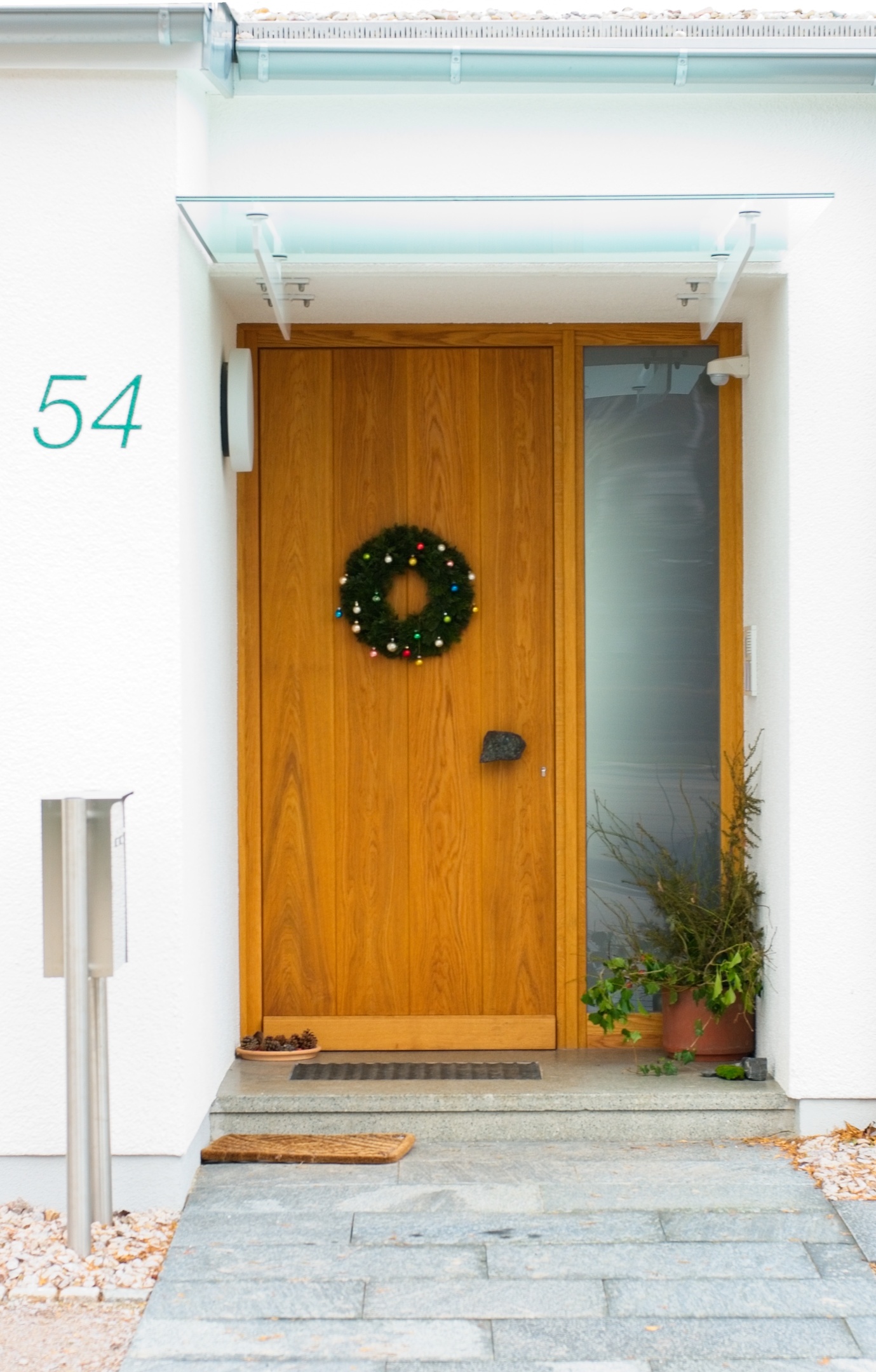 Ein Weg aus Granitplatten, endet vor einer Haustür aus hellem Holz. An der rechten Seite ein schmales Milchglasfenster. Mit Glasdach überdacht.
