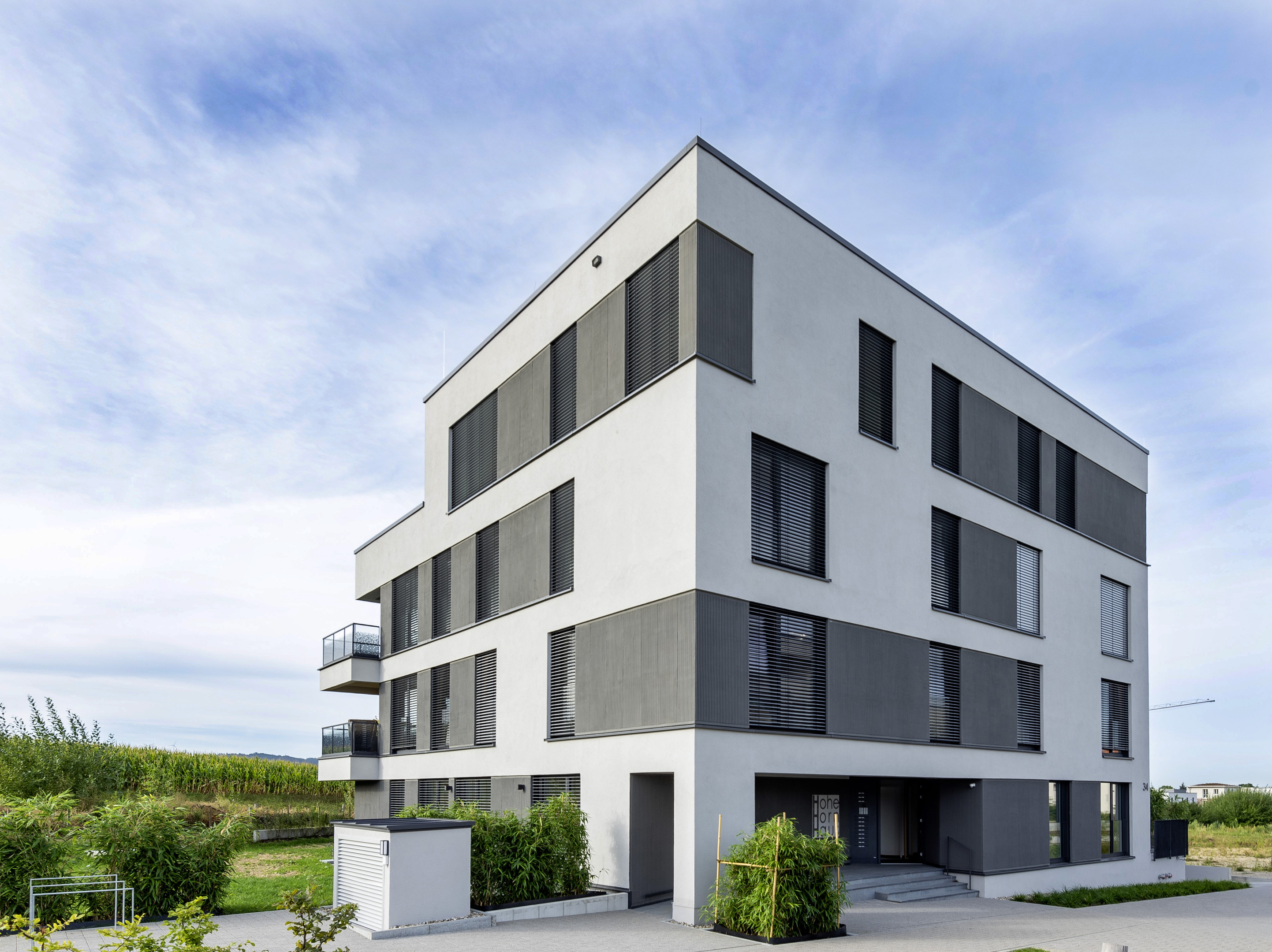 Ein würfelförmiges, vierstöckiges Haus. Der Hauseingang ist zurückgesetzt. Im obersten Stockwerk eine Dachterrasse, darunter auskragende Balkone.
