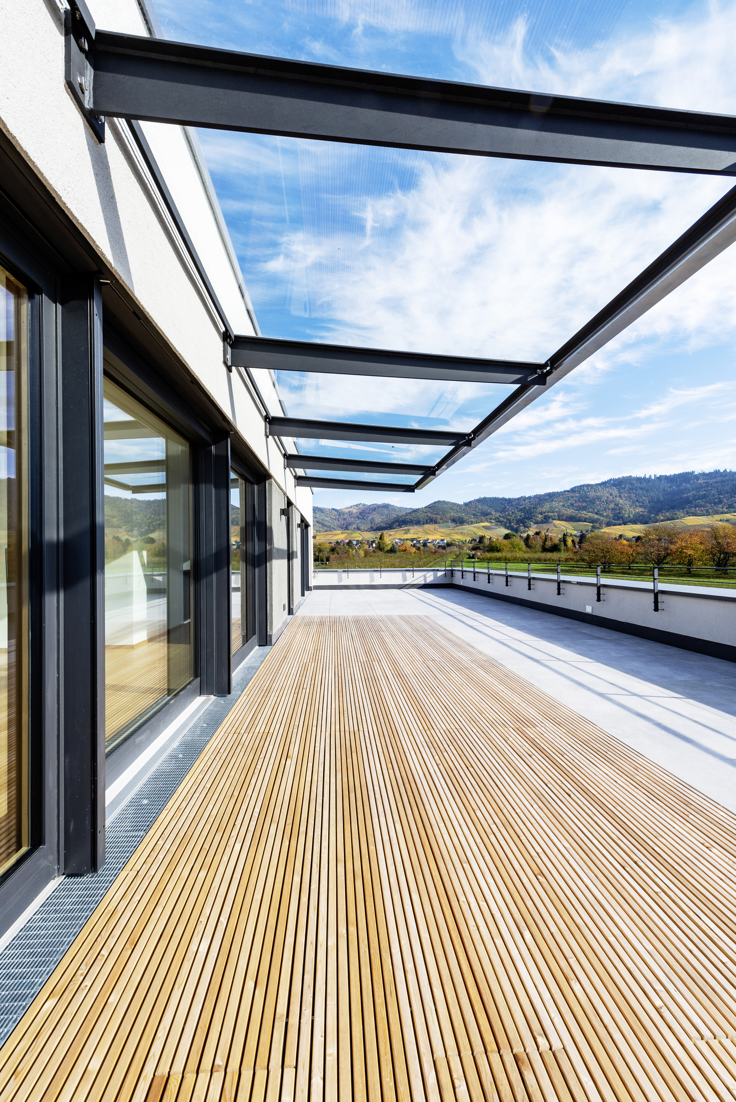 Dachterrasse, die links von einer Fensterfront begrenzt ist. Unter einem langen Vordach aus Glas ein Bodenbelag aus schmalen parallelen Holzlamellen.