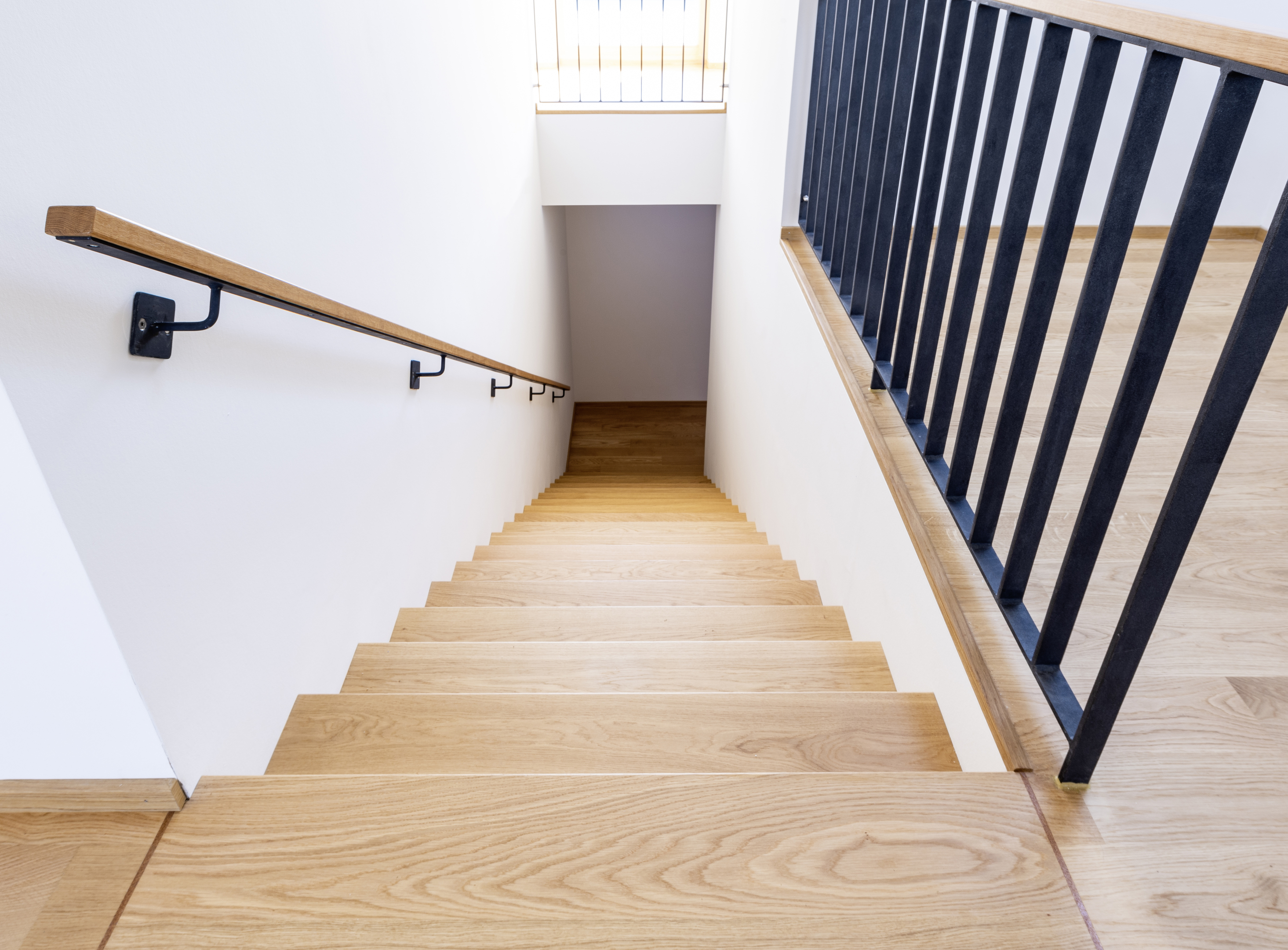 Eine Treppe mit Holzbelag, die zwischen weißen Wänden nach unten führt. Der Handlauf ist aus schwarzem Metall mit Holzauflage.